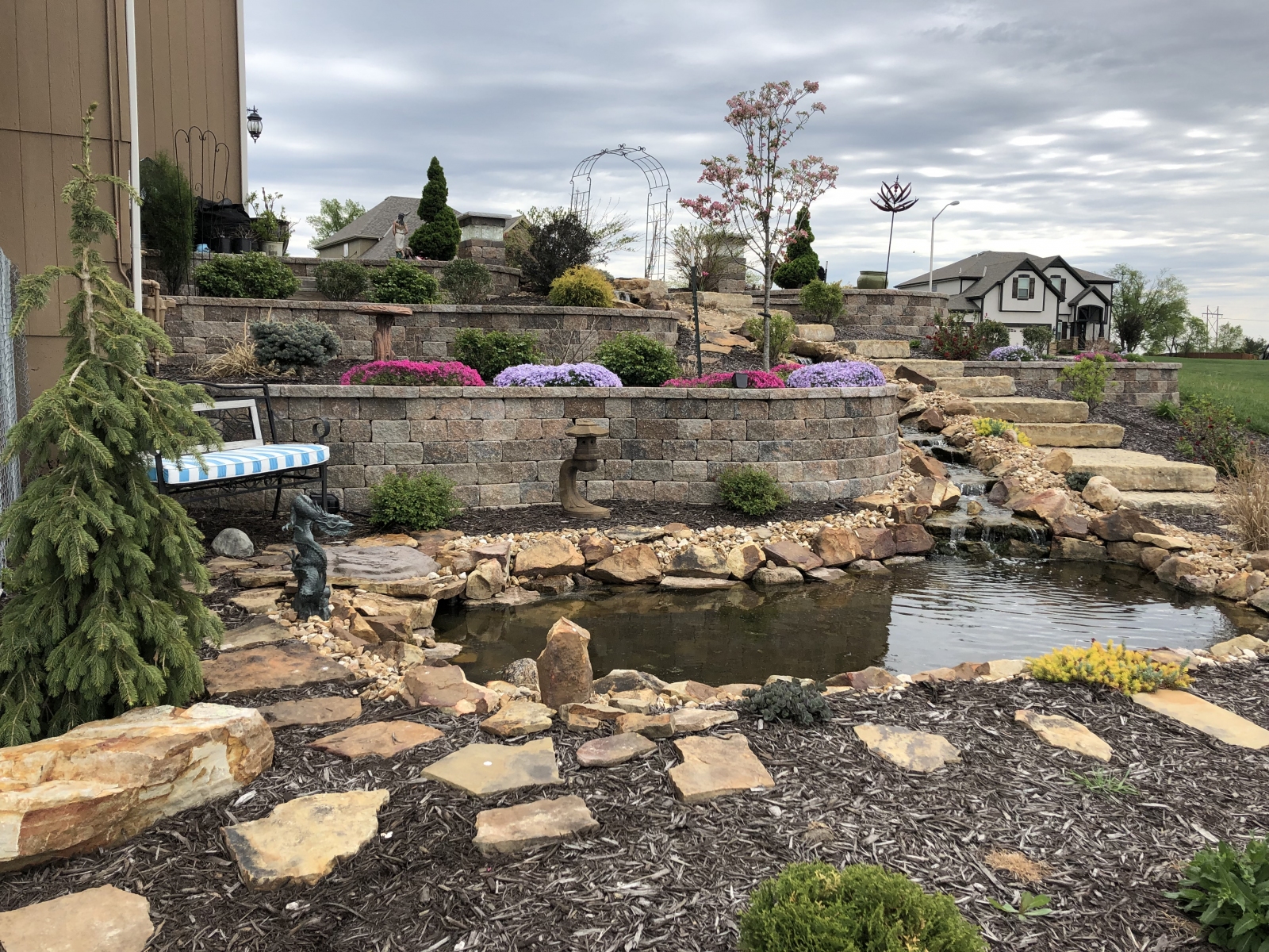 A photo of a landscaping project that uses a variety of sizes and types of stone to create tiered bedding areas, a flowing stream and a decorative pool at the bottom.