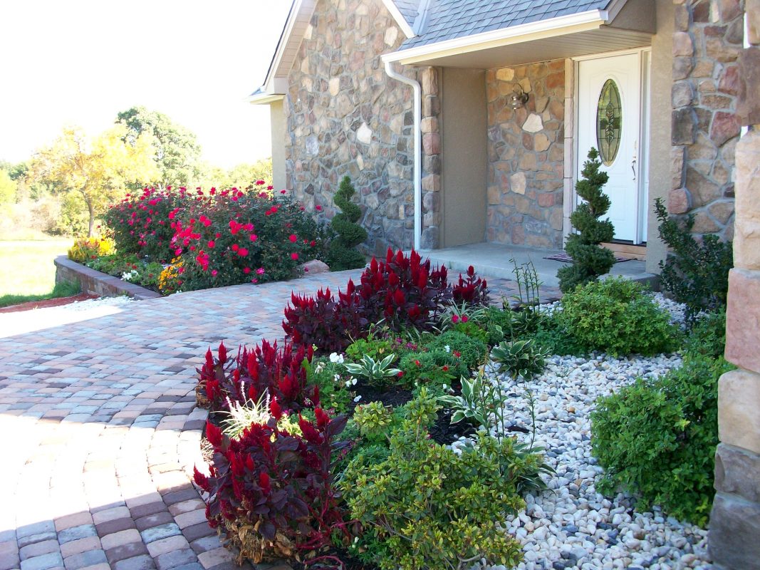 A photo of a brick sidewalk approach to a stone house with a rock bedding area of various plants.