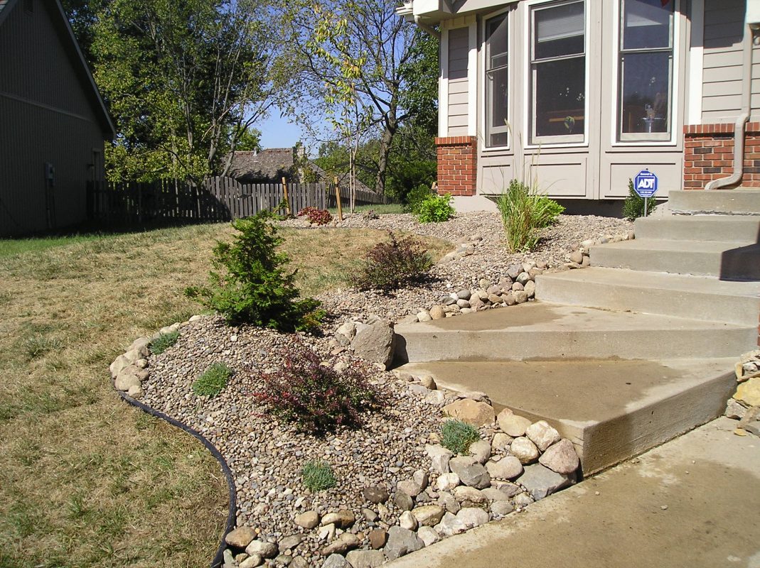 A photo of the front corner of a home with steps leading to the front door. To the left of the steps is a scalloped bedding area using a variety of stone sizes and types as the base with various live plants. 