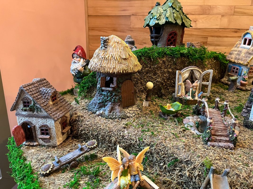 Indoor Fairy Garden brand display, includes a teeter totter, boyo and girl fairies sitting on a bench together, a wooden bridge, a grome, fairy houses of various sizes and shapes, and other small accessories.