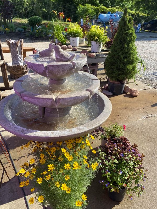 Three-tiered fountain with love birds on top tier. Displayed on concrete pad with potted plants at base. Background includes potted trees, plants, large propane tank and large trees in far background.