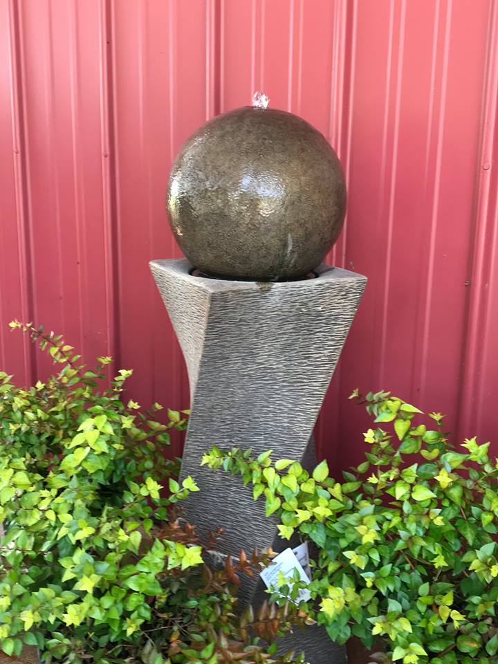 Bubbler fountain with large copper-colored ball perched on square twisted base. Potted shrubs surround base of fountain. On display at Full Features Garden Center.
