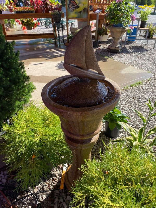 Modern fountain with sailboat figure on top. Displayed on gravel pad at Full Features Garden Center, with potted plants surrounding fountain base. Background includes various plants and pots.