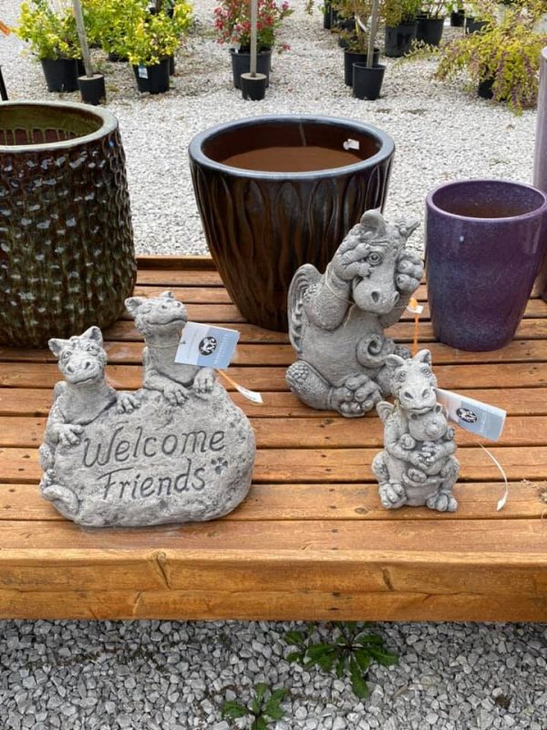 Dragon statuary on wooden display shelf at Full Features Garden Center. Statue 1 - two small dragons behind a rock that says "welcome friends." Statue 2 - Mama dragon cuddling baby dragon. Statue 3 - seated dragon peeking through his fingers as if hiding. Various pots and plants in background.