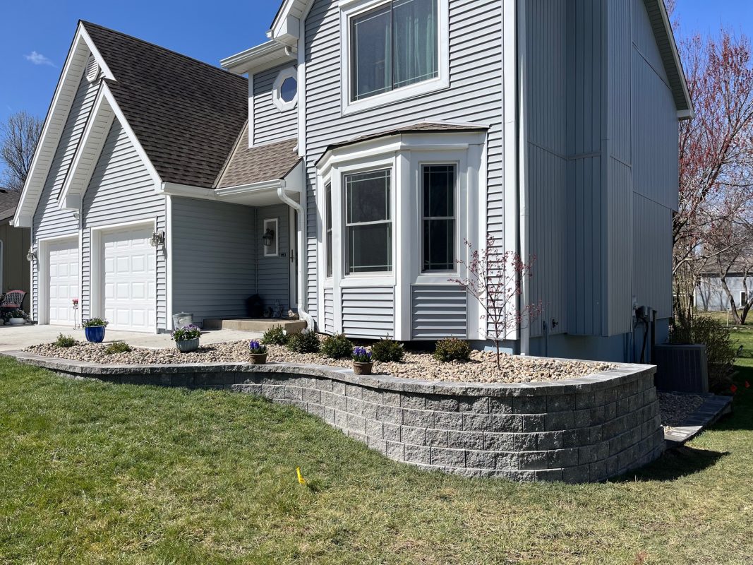 Gray house with white trim, custom landscape project using block retaining walls, featuring shrubs and plants in rock gardens.